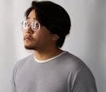 A thoughtful man wearing glasses and a striped shirt, looking to the side in a studio setting.
